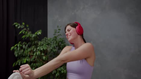 Close-up-of-a-happy-and-joyful-athletic-brunette-girl-wearing-a-purple-top-and-red-wireless-headphones-dancing-and-having-fun-while-listening-to-music-after-playing-sports-in-a-modern-apartment