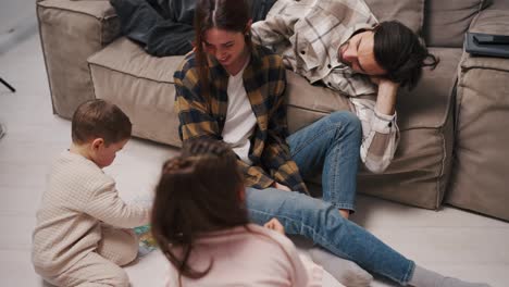 A-happy-brunette-girl-in-a-plaid-shirt-and-blue-jeans-sits-on-the-floor-and-plays-with-her-children-her-husband-lies-near-them-on-the-sofa-they-spend-fun-time-together-in-a-modern-studio-apartment