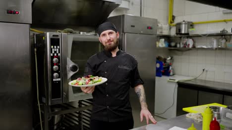 Retrato-De-Un-Chef-Masculino-Seguro-Y-Feliz-Con-Un-Uniforme-Negro-Y-Barba-Que-Sostiene-Su-Ensalada-Preparada-En-Sus-Manos-Antes-De-Servirla-En-Un-Restaurante-En-La-Cocina.