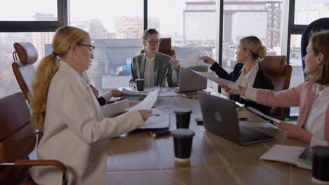 A-group-of-businesswomen-in-business-clothes-are-communicating-during-a-meeting-and-passing-each-other-sheets-with-graphs-and-ideas-about-their-future-work-in-a-modern-society-with-large-windows