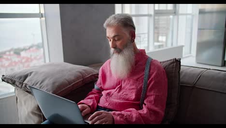 Porträt-Eines-älteren-Mannes-Mit-Grauem-Haar-Und-üppigem-Bart-In-Einem-Rosa-Hemd,-Der-Auf-Einem-Braunen-Sofa-In-Einer-Modernen-Wohnung-Sitzt-Und-Auf-Einem-Grauen-Laptop-Tippt