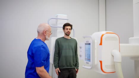 A-confident-experienced-male-doctor-with-glasses-in-a-blue-uniform-makes-a-fluorography-of-a-brunette-guy-in-a-green-jacket-during-an-appointment-in-the-office-in-a-modern-clinic