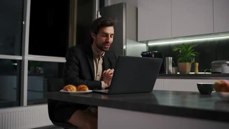 A-serious-and-confident-brunette-man-with-stubble-in-a-black-jacket-and-beige-shorts-goes-on-a-video-call-using-a-laptop-for-a-video-conference-in-the-evening-working-remotely-in-a-modern-apartment