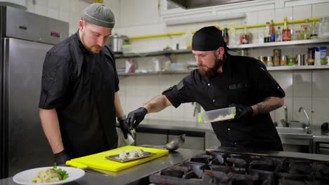 Confident-male-cook-in-a-black-uniform-and-bandana-sprinkles-a-finished-dish-with-cheese-in-the-kitchen.-Duo-of-chefs-in-the-kitchen-preparing-a-dish-together