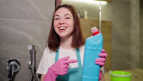 Portrait-of-a-joyful-and-confident-brunette-cleaning-lady-in-a-white-T-shirt-and-a-blue-apron-who-points-to-the-detergent-in-her-hand-and-gives-it-a-like-in-a-pink-rubber-glove-in-the-bathroom-of-a-modern-apartment-while-cleaning