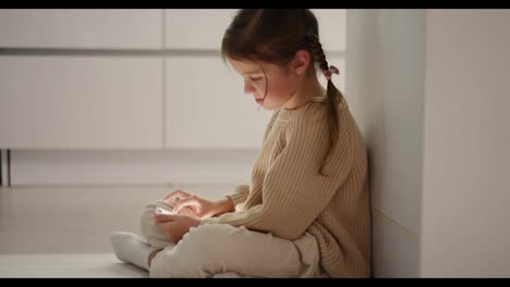 Close-up-of-a-little-brown-haired-girl-in-a-cream-sweater-sitting-on-the-floor-near-the-kitchen-table-and-doing-something-on-the-phone-during-her-time-alone-in-the-evening