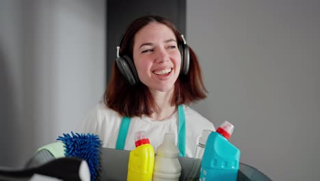 A-cheerful-brunette-cleaning-lady-listens-to-music-in-black-wireless-headphones-walks-along-the-apartment-corridor-and-carries-a-gray-plastic-basin-with-cleaning-tools-and-detergents