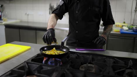 Confident-male-chef-in-a-black-uniform-and-with-black-protective-gloves-pours-alcohol-onto-fried-vegetables-in-a-frying-pan-to-apply-the-flambing-technique-in-a-modern-kitchen-in-a-restaurant