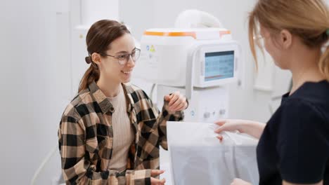 A-happy-brunette-girl-in-round-glasses-in-a-plaid-shirt-receives-the-results-of-her-fluorography-in-pictures-in-a-special-package-from-a-girl-doctor-in-the-fluorography-room-in-a-modern-clinic