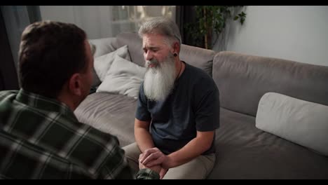 Over-the-shoulder-A-sad-man-with-gray-hair-and-a-lush-gray-beard-in-a-gray-T-shirt-communicates-with-his-boyfriend-a-brunette-with-stubble-in-a-checkered-shirt-and-holds-his-hands-while-sitting-on-a-gray-sofa-during-a-difficult-conversation-among-men-in-an-LGBT-couple-in-the-evening-in-a-modern-apar