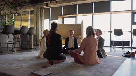 Group-of-confident-businesswomen-in-business-suits-meditate-while-sitting-on-a-carpet-in-a-modern-office.-Confident-business-girls-sitting-on-a-gray-soft-carpet-and-meditating-during-a-break-between-work-in-the-office