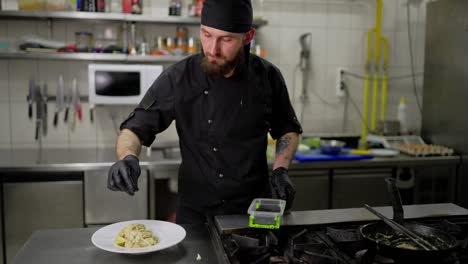 Un-Hombre-Con-Barba,-Un-Chef-Profesional-Con-Un-Uniforme-Protector-Negro,-Rocía-Un-Plato-Con-Especias-Y-Hierbas-Mientras-Lo-Prepara-Para-Servir-En-La-Cocina.