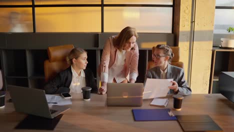 A-trio-of-confident-businesswomen-in-business-clothes-and-jackets-gathered-around-a-wooden-table-in-an-office-and-discussing-the-current-plan-and-solution-to-a-problem-using-sheets-and-a-laptop-in-a-sunny-office