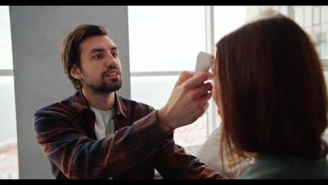 Un-Chico-Moreno-Con-Barba-Incipiente,-Con-Una-Camisa-A-Cuadros-Rojos-Y-Azules-Y-Una-Camiseta-Blanca,-Mide-La-Temperatura-Con-Un-Termómetro-Electrónico-De-Su-Novia-Morena-Con-Un-Suéter-Verde-Mientras-Está-Sentada-En-El-Sofá-De-Un-Moderno-Apartamento-Con-Grandes-Ventanales.