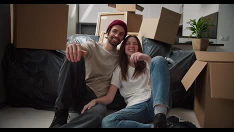 Portrait-of-a-happy-brunette-man-with-stubble-in-a-red-hat-and-a-beige-T-shirt-who-sits-on-the-floor-with-his-brunette-girlfriend-in-a-white-T-shirt-near-the-sofa-in-a-black-cellophane-case-among-a-pile-of-boxes-in-a-new-house-after-a-housewarming-party