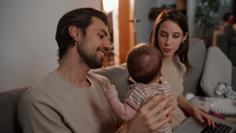 Happy-brunette-man-with-stubble-holds-his-little-baby-daughter-in-his-arms-along-with-his-young-wife-in-a-modern-apartment-on-a-gray-sofa