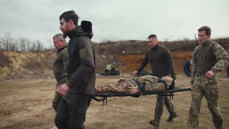 Side-view-of-four-confident-male-soldiers-in-dark-green-uniforms-carrying-a-wounded-doll-of-a-male-soldier-in-camouflage-uniform-on-a-stretcher-and-placing-him-on-the-ground-near-an-ambulance-on-a-training-ground-during-a-military-medicine-exercise