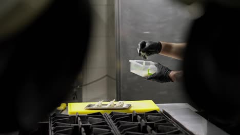 Side-view-of-a-confident-professional-chef-in-a-black-uniform-sprinkles-a-finished-dish-with-cheese-while-cooking-in-the-kitchen-in-a-restaurant