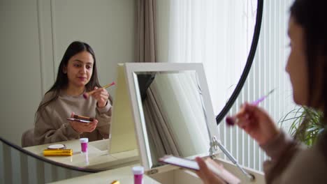 Glückliches-Brünettes-Mädchen-Mit-Grauem-Pullover-Beim-Schminken-Vor-Dem-Spiegel-In-Einem-Modernen-Apartment.-Glückliches-Mädchen-Mit-Brauner-Haut-Macht-Make-up-Und-Trägt-Tagsüber-In-Einem-Hellen,-Modernen-Raum-Puder-Auf-Ihr-Gesicht-Auf