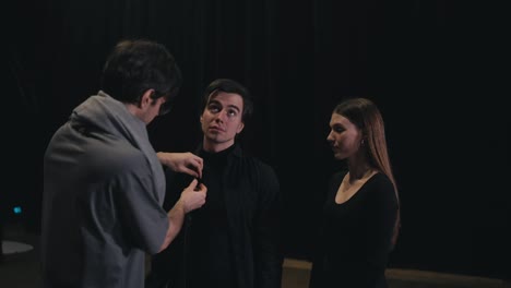 A-confident-brunette-man-in-a-gray-T-shirt-and-scarf-the-stage-director-adjusts-the-black-costumes-of-the-actors-before-they-go-on-stage-during-a-performance-in-the-theater