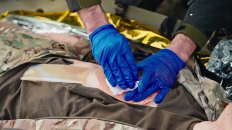 Primer-Plano-De-Un-Médico-Militar-Confiado-Con-Guantes-Médicos-De-Goma-Azules-En-Sus-Manos-Endereza-El-Pecho-De-Un-Soldado-Herido-Y-Detiene-El-Sangrado-Usando-Un-Yeso-Especial-Mientras-Brinda-Primeros-Auxilios-En-Combate.