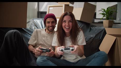 A-happy-brunette-girl-in-a-white-T-shirt-plays-with-her-boyfriend-with-stubble-in-a-video-game-using-two-joysticks-near-them-there-are-a-large-number-of-boxes-and-a-sofa-in-a-black-cover-after-moving