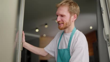 Close-up-a-blond-janitor-guy-in-a-white-T-shirt-and-blue-apron-washes-glass-walls-in-a-modern-apartment-during-cleaning