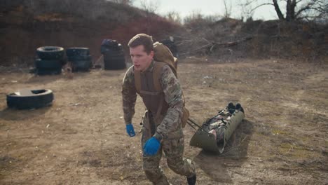 A-tired-confident-young-military-man-in-a-camouflage-uniform-and-blue-medical-gloves-drags-an-unconscious-soldier-in-army-camouflage-with-the-help-of-a-special-stretcher-in-the-steppe