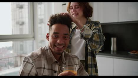 Portrait-of-a-cheerful-and-happy-young-man-with-Black-brunette-skin-color-in-a-checkered-beige-shirt-who-sits-and-drinks-juice-while-his-girlfriend-touches-his-hairstyle-in-a-modern-apartment