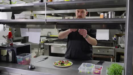 A-confident-male-cook-in-a-black-uniform-stands-in-the-kitchen-while-issuing-orders-lays-out-stickers-with-written-dishes-and-gives-out-the-required-dish-to-the-hall-for-the-waiters-to-hand-over-to-customers-in-the-restaurant