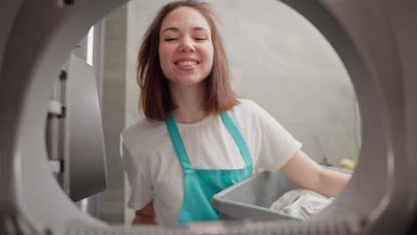 A-happy-brunette-girl-in-a-white-T-shirt-and-a-blue-apron-took-out-clean-things-from-the-washing-machine-and-closes-its-door-while-cleaning-the-apartment-on-call