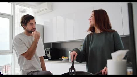 Una-Chica-Morena-Feliz-Con-Un-Suéter-Verde-Prepara-El-Desayuno-Mientras-Su-Novio,-Un-Hombre-Moreno-Con-Un-Escudo-Y-Una-Camiseta-Blanca,-Se-Apura-Los-Dientes-En-La-Cocina-De-Un-Apartamento-Moderno.