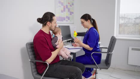 A-confident-brunette-pediatrician-girl-in-a-blue-uniform-communicates-with-a-young-father-and-gives-him-advice-during-an-appointment-with-a-childrens-doctor-and-pediatrician-in-a-modern-clinic