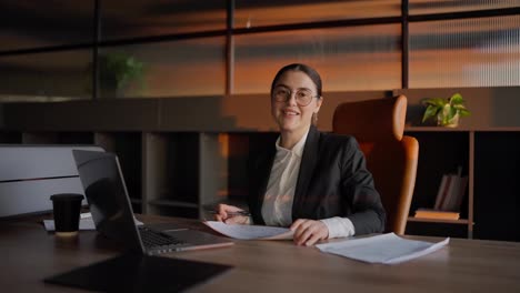 Retrato-De-Una-Empresaria-Morena-Feliz-Con-Gafas-Redondas-Y-Uniforme-De-Negocios-Sentada-En-Una-Mesa-Frente-A-Una-Computadora-Portátil-Y-Trabajando-En-Una-Oficina-Soleada