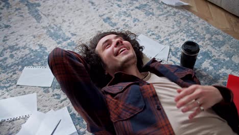 Happy-brunette-guy-with-curly-hair-in-a-checkered-shirt-lies-on-a-pillow-on-the-floor-surrounded-by-sheets-of-paper-and-a-cup-of-coffee-at-home-while-working