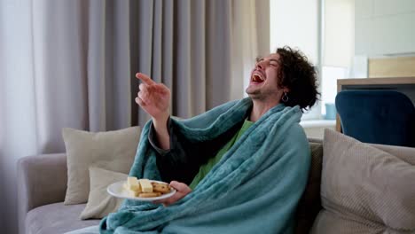 Ein-Fröhlicher-Und-Freudiger-Brünetter-Typ-Mit-Lockigem-Haar-Hält-Süße-Snacks-In-Den-Händen-Und-Lacht,-Während-Er-Sich-Zu-Hause-Auf-Der-Couch-Eine-Lustige-Komödie-Ansieht,-In-Eine-Decke-Gehüllt