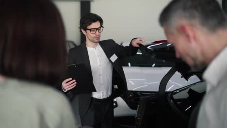 Over-the-shoulder-A-confident-brunette-guy-with-glasses-and-a-business-suit-with-a-tablet-in-his-hands-shows-his-family-the-trunk-of-a-modern-car-in-a-car-showroom