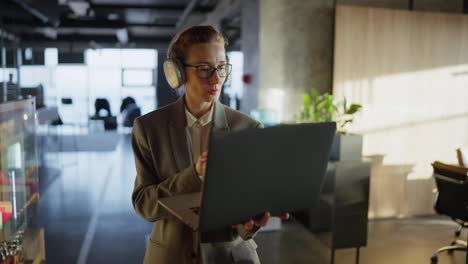 A-confident-middle-aged-blonde-girl-with-glasses-in-a-gray-business-suit-walks-through-a-modern-office-and-types-on-her-laptop-while-working-in-a-sunny-office