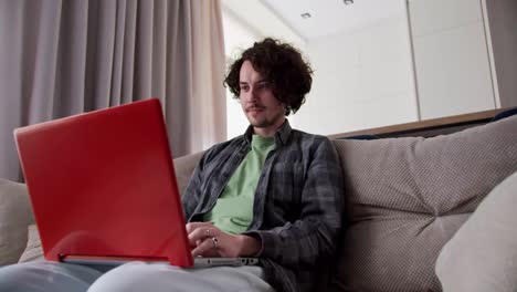 Zoom-in-happy-brunette-guy-with-curly-hair-and-mustache-in-a-checkered-shirt-is-very-happy-about-his-success-and-Victory-while-working-on-a-red-laptop-on-the-sofa-at-home