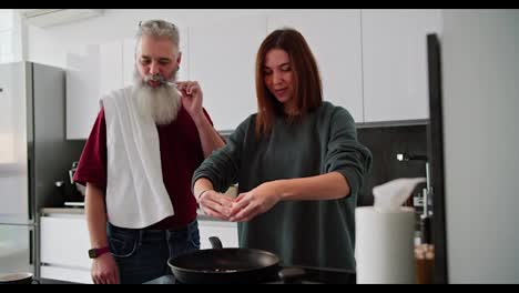 Happy-elderly-man-with-gray-hair-and-lush-beard-brushes-his-teeth-with-a-towel-on-his-shoulders-while-his-adult-brunette-daughter-in-a-green-sweater-makes-breakfast-in-a-modern-kitchen