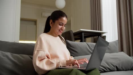 Happy-brunette-girl-in-a-pink-jacket-and-green-pants-sits-on-a-gray-sofa-and-types-on-her-laptop-while-working-remotely-at-home-happy-during-the-day