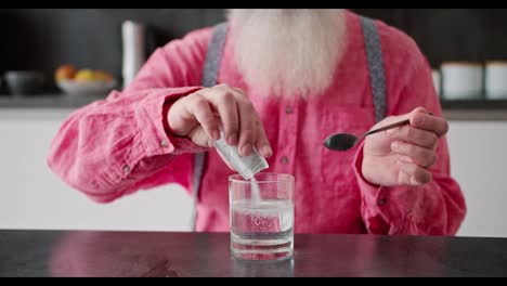 Nahaufnahme-Eines-Mannes-Mit-Grauem-Haar-Und-üppigem-Bart-In-Einem-Rosa-Hemd,-Der-Weiße-Medizin-In-Ein-Transparentes-Glas-Wasser-Für-Sich-Selbst-Gießt-Und-Es-In-Einer-Modernen-Wohnung-Umrührt