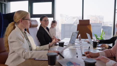 Confident-brunette-girl-in-business-clothes-holds-a-small-infant-baby-in-her-arms-while-taking-part-in-a-business-meeting-at-a-table-in-a-modern-office-with-large-windows