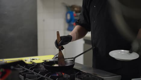 Side-view-of-a-professional-chef-in-a-black-uniform-transferring-vegetables-and-seafood-to-a-hot-frying-pan-while-preparing-food-on-a-gas-burner-in-a-restaurant-kitchen