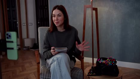 Confident-brunette-girl-communicates-with-her-viewers-while-blogging-and-sits-on-a-chair-and-takes-notes-using-a-notepad-in-a-modern-cozy-apartment