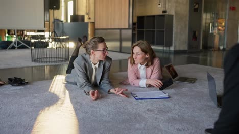 Middle-aged-blondes-in-business-clothes-lie-on-the-floor-on-a-gray-soft-carpet-and-communicate-during-a-break-at-work.-In-the-office