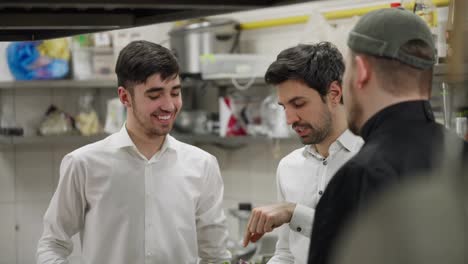 Los-Camareros-Con-Camisas-Blancas-Se-Comunican-Con-Un-Chef-Profesional-Con-Camisa-Negra-Sobre-Un-Plato-Específico-En-La-Cocina-De-Un-Restaurante.