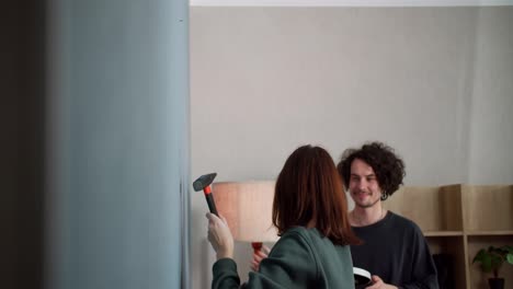 Confident-brunette-girl-in-a-green-jacket-hits-a-nail-with-a-hammer-and-hangs-a-clock-on-the-wall-while-spending-time-with-her-boyfriend-at-home