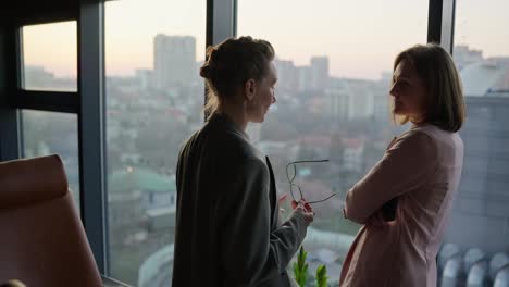 Two-middle-aged-businesswomen-in-business-uniforms-stand-near-panoramic-windows-in-a-modern-office-and-communicate-about-current-issues