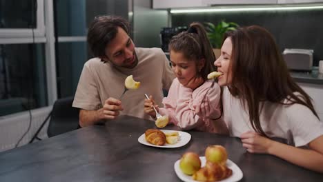 A-happy-brunette-man-with-stubble-in-a-beige-T-shirt-together-with-his-brunette-wife-in-a-white-T-shirt-and-a-little-daughter-in-a-pink-suit-eat-cheesecakes-during-their-breakfast-in-a-modern-apartment-at-a-black-dining-table-in-the-kitchen-in-the-morning